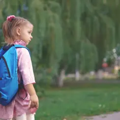 Mała dziewczynka z niebieskim plecakiem, z kwiatkiem we włosach, ma na twarzy oznaki niezadowolenia, być może buntu przed pójściem do szkoły.