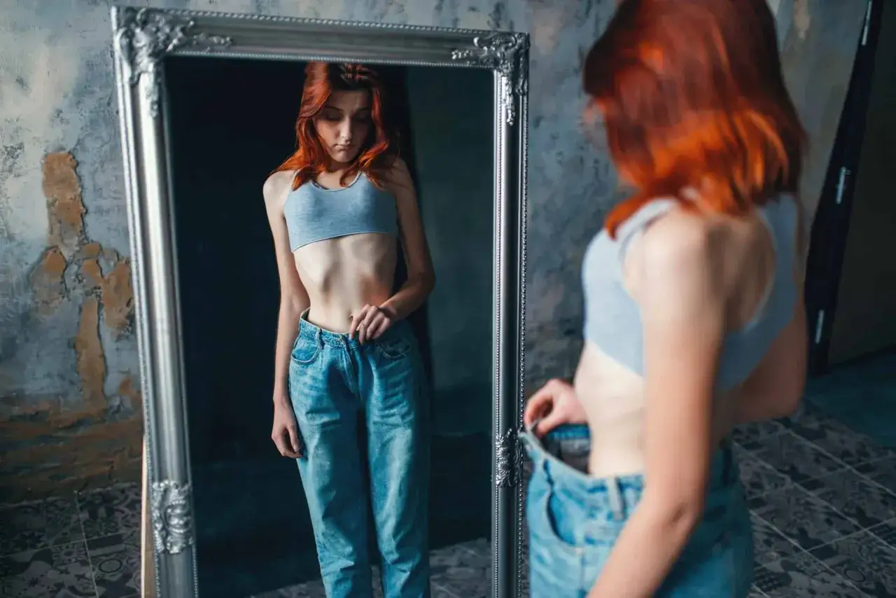 Dziewczyna z rudymi włosami patrzy w lustro, widząc swoje wychudzone ciało. Jej twarz wyraża smutek, odzwierciedlając body dysphoria.