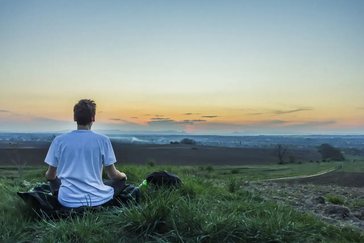 Chłopak w białej koszulce siedzi na kocu na wzgórzu, medytując. Zachód słońca tworzy piękne tło. To chwila mindfulness.