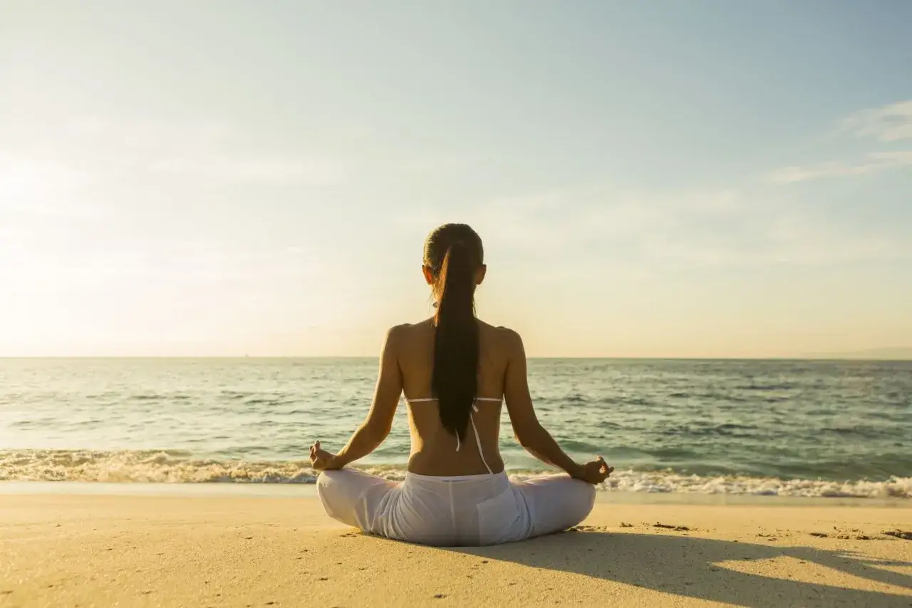 Kobieta w pozycji lotosu na plaży, praktykująca mindfulness. Słońce zachodzi nad oceanem, tworząc spokojną atmosferę.